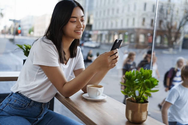 Schönes asiatisches Mädchen, das auf Handy, während im Stadtcafé nahe Fenster sitzt, Kaffee trinkt und Smartphone verwendet.