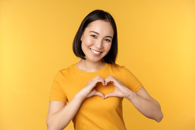Schönes asiatisches Mädchen, das Herzliebesgeste und lächelnde weiße Zähne zeigt, drückt Fürsorge und Sympathie aus, die über gelbem Hintergrund stehen