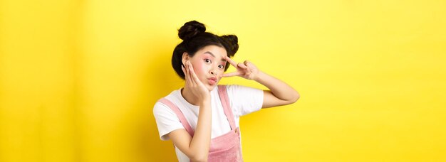 Schönes asiatisches Mädchen, das Vsign zeigt und süß schmollend macht, dummes Gesicht mit Make-up, das auf Gelb steht