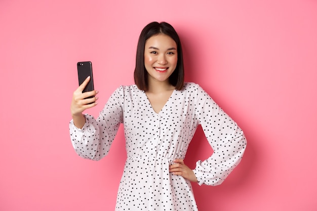 Schönes asiatisches Mädchen mit Fotofilter-App und Selfie auf dem Handy, posiert in süßem Kleid vor rosa Hintergrund.