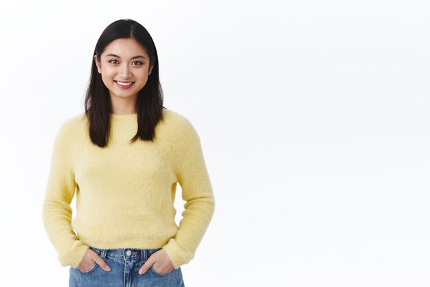 Schönes asiatisches Mädchen mit glühender Haut und weißen Zähnen lächelnd glücklich Händchen haltend in Jeanstaschen lässige Pose vor weißem Hintergrund in der Nähe einer leeren Werbefläche, die das Produkt vorstellt