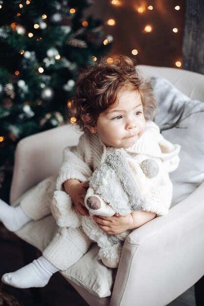 Schönes Baby mit lockigem Haar in Weiß, das im Stuhl mit Teddybär sitzt.