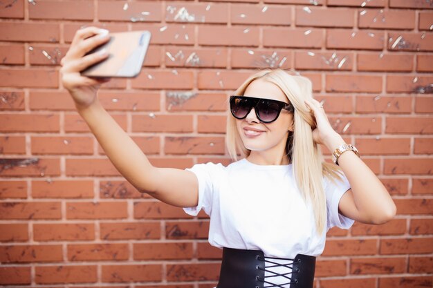 Schönes blondes Frauenmädchen, das ein selfie auf Smartphone nimmt, das gegen rote Backsteinmauer stehend aufwirft.