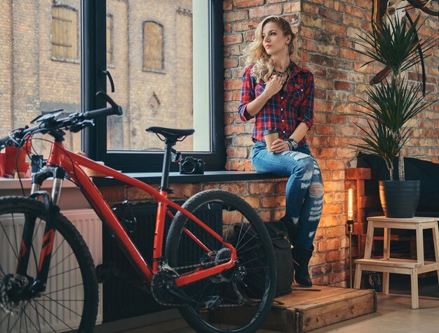 Schönes blondes Hipster-Mädchen mit langen lockigen Haaren, gekleidet in ein Fleece-Shirt und Jeans, hält eine Tasse Kaffee zum Mitnehmen auf einer Fensterbank in einem Studio mit Loft-Interieur.