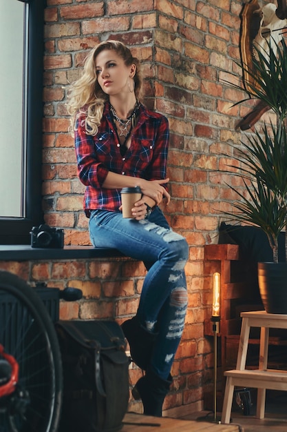 Schönes blondes Hipster-Mädchen mit langen lockigen Haaren, gekleidet in ein Fleece-Shirt und Jeans, hält eine Tasse Kaffee zum Mitnehmen auf einer Fensterbank in einem Studio mit Loft-Interieur.