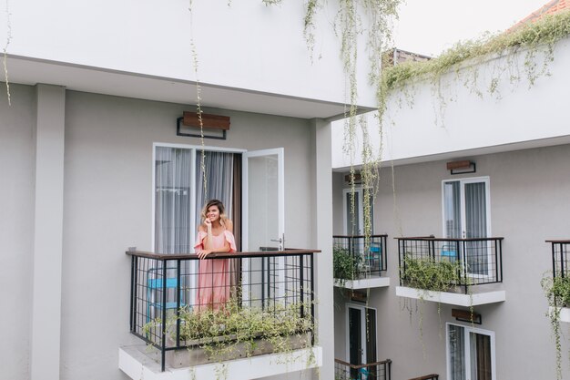 Schönes blondes Mädchen, das am Hotelbalkon aufwirft. lächelnde romantische Frau im rosa Kleid, die Morgen genießt.