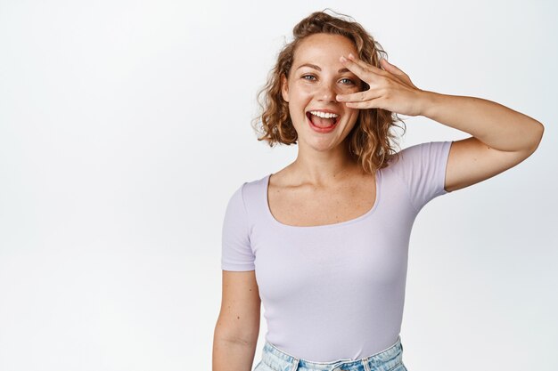 Schönes blondes Mädchen, das Augenfriedensgeste zeigt, sorglos lacht und lächelt und im T-Shirt auf Weiß steht. Platz kopieren