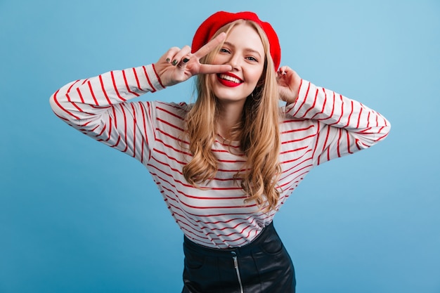 Schönes blondes Mädchen, das Friedenszeichen auf blauer Wand zeigt. freudiges französisches weibliches Model.