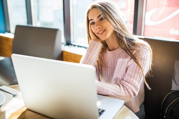 Schönes blondes Mädchen im Modeweißpullover bei der Arbeit an ihrem Computer im Café in der täglichen Zeit