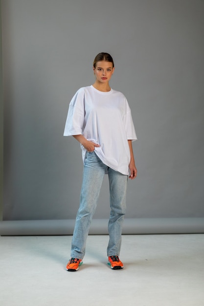 Schönes blondes Mädchen in einem weißen übergroßen T-Shirt und blauen Jeans posiert auf einem grauen Hintergrund