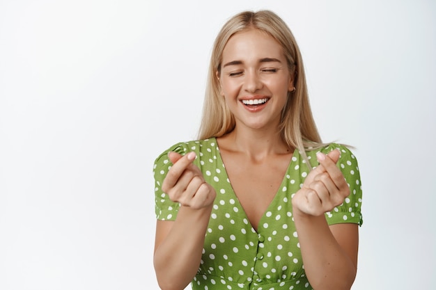 Schönes blondes Mädchen mit gebräunter Haut, zeigt Fingerherzen und lächelt glücklich, steht auf Weiß.