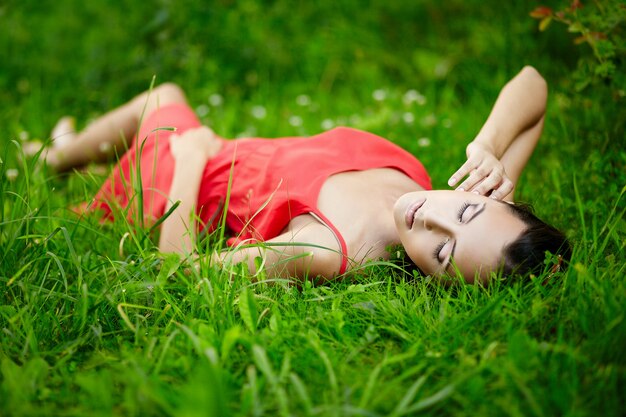 schönes brünettes Frauenmodell, das im grünen Sommerhellgras im Park mit Make-up im roten Kleid liegt.