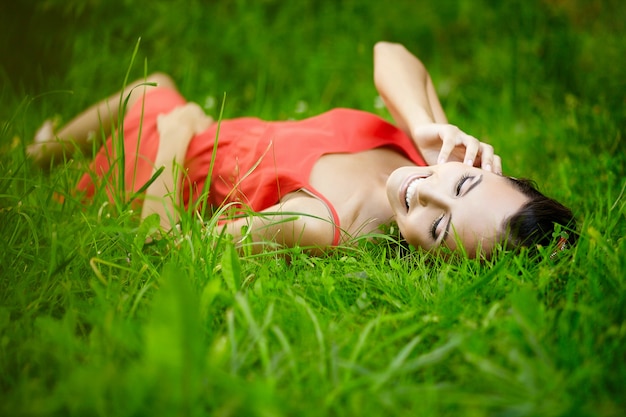 schönes brünettes Frauenmodell, das im grünen Sommerhellgras im Park mit Make-up im roten Kleid liegt.