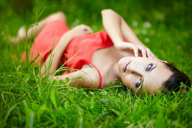 schönes brünettes Frauenmodell, das im grünen Sommerhellgras im Park mit Make-up im roten Kleid liegt.