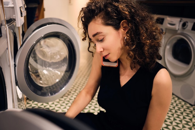 Schönes brünettes Mädchen, das auf dem Smartphone spricht und Wäsche in modernen Selbstbedienungswäschereien wäscht