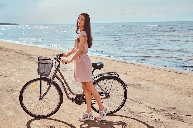 Schönes brünettes Mädchen in Kleid und Hut, das an einem sonnigen Tag mit einem Fahrrad am Strand posiert