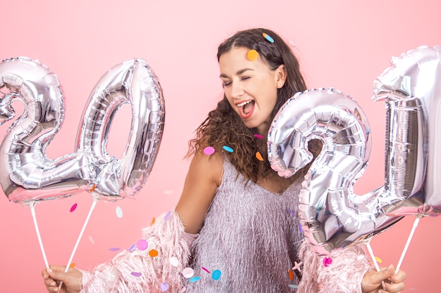 Schönes brünettes Partygirl mit lockigem Haar und festlichen Kleidern, die auf einem rosa Studiohintergrund mit Konfetti aufwerfen und silberne Luftballons für das Neujahrskonzept halten