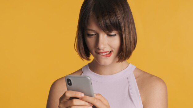 Schönes charmantes brünettes Mädchen mit Bob-Haar beißenden Lippen, die mit ihrem Freund auf dem Smartphone über gelbem Hintergrund plaudern