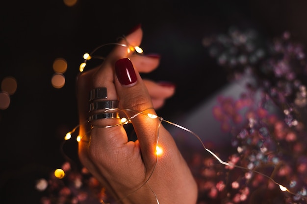 Schönes dunkles Foto der Handfinger der Frau mit großem silbernem Ring auf Blumen und leuchtenden Lichtern
