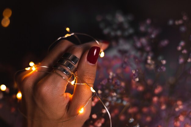 Schönes dunkles Foto der Handfinger der Frau mit großem silbernem Ring auf Blumen und leuchtenden Lichtern