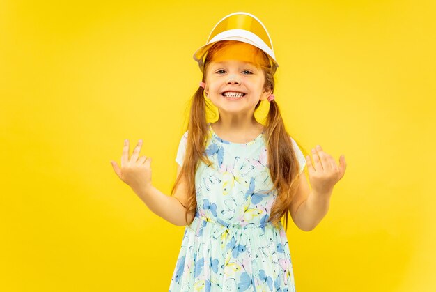 Schönes emotionales kleines Mädchen lokalisiert auf gelbem Hintergrund. Halblanges Porträt eines glücklichen und feiernden Kindes, das ein Kleid und eine orangefarbene Mütze trägt. Konzept des Sommers, der menschlichen Gefühle, der Kindheit.