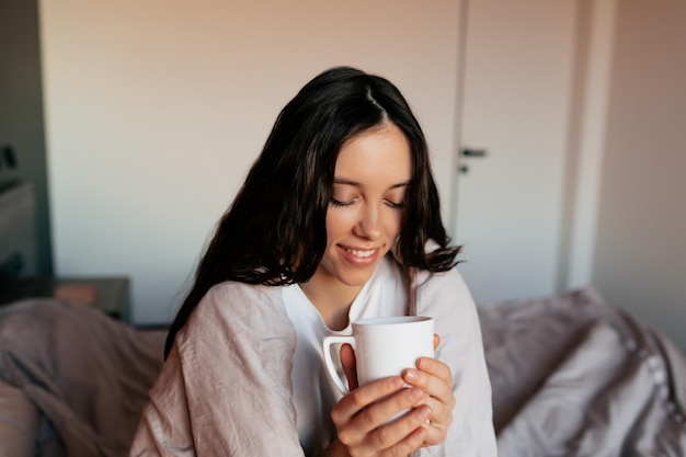 Schönes entzückendes Mädchen mit dunklem Haar hält eine Tasse Kaffee und deckt morgens bei Sonnenschein eine Decke im Schlafzimmer ab Home-Konzept Morgens aufwachen zu Hause