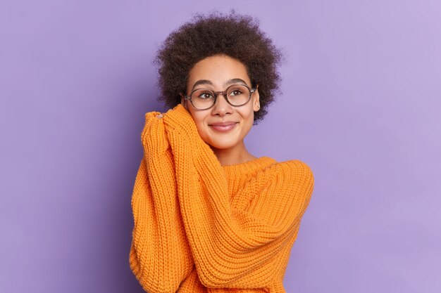 Schönes ethnisches Mädchen mit natürlichem Afro-Haar hält Hände in der Nähe des Gesichts und sieht mit verträumtem Ausdruck aus, trägt irgendwo einen losen orangefarbenen Strickpullover.