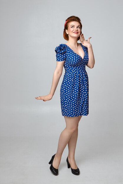 Schönes europäisches Pin-up-Girl in voller Länge in stilvollem blauem Kleid mit tiefem Ausschnitt und schüchternem Aussehen, breit lächelnd, während es mit jemandem flirtet. Menschliche Mimik und Körper