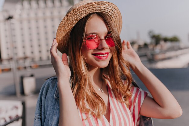 Schönes freudiges Mädchen in der rosa Brille, die mit sanftem Lächeln aufwirft. Außenaufnahme der stilvollen weißen Frau, die ihren Strohhut auf Stadt berührt.