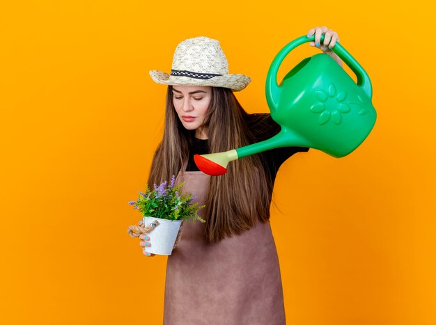 Schönes Gärtnermädchen, das Uniform und Gartenhut trägt, die Blume im Blumentopf mit Bewässerungsdose lokalisiert auf orange Hintergrund tränkt