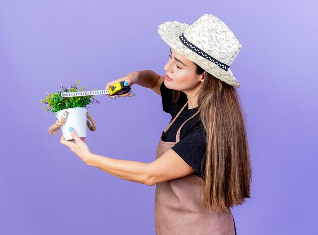 Schönes Gärtnermädchen in der Uniform, die Gartenhut misst Blume im Blumentopf mit Maßband lokalisiert auf blauem Hintergrund