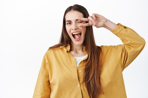 Schönes glückliches Mädchen zeigt ein Friedens-V-Zeichen über dem Auge, zwinkert und lächelt breit, drückt einen positiven und fröhlichen Lebensstil aus und steht in stilvollem Outfit gegen weiße Wand