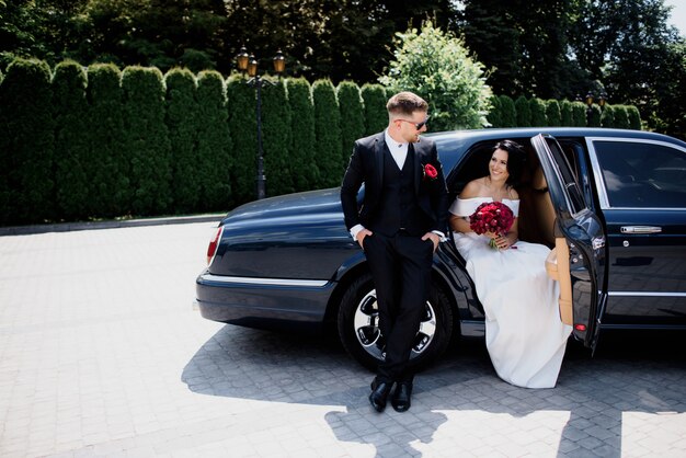 Schönes Hochzeitspaar lächelt im schwarzen Auto am sonnigen Tag, gekleidet in eleganten Hochzeitsoutfits mit rotem Blumenstrauß