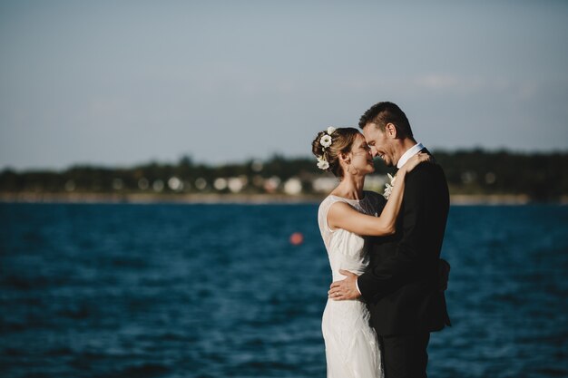 Schönes Hochzeitspaar umarmt sich die zarte Stellung, die auf dem Seeufer steht