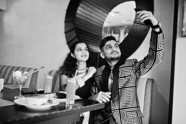 Schönes indisches verliebtes Paar trägt Saree und eleganten Anzug, sitzt im Restaurant und macht Selfie per Telefon zusammen Auf dem Tisch Dessert Kuchen Eis und Saft