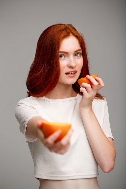 schönes Ingwermädchen, das Orangen über graue Wand hält.