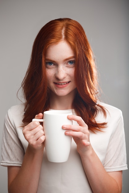 schönes Ingwermädchen, das Tasse über graue Wand mit Gegenlicht hält.