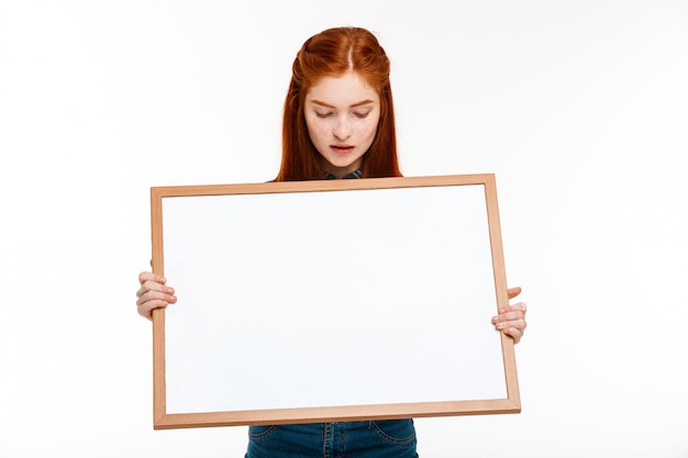 schönes Ingwermädchen mit Whiteboard über weißer Wand.