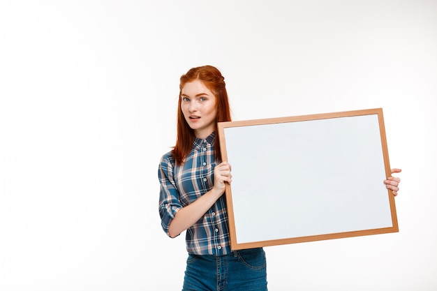 schönes Ingwermädchen mit Whiteboard über weißer Wand.