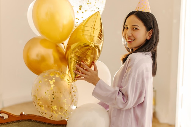 Schönes junges asiatisches brünettes Mädchen, das drinnen mit Luftballons in die Kamera posiert Feiertagsfeierkonzept