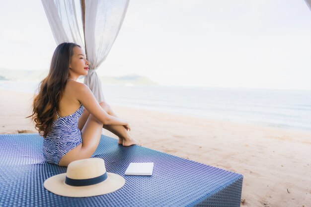 Schönes junges asiatisches Frauenlesebuch des Porträts mit glücklichem Lächeln entspannen sich im Klubsessel auf dem Strandseeozean für Freizeit