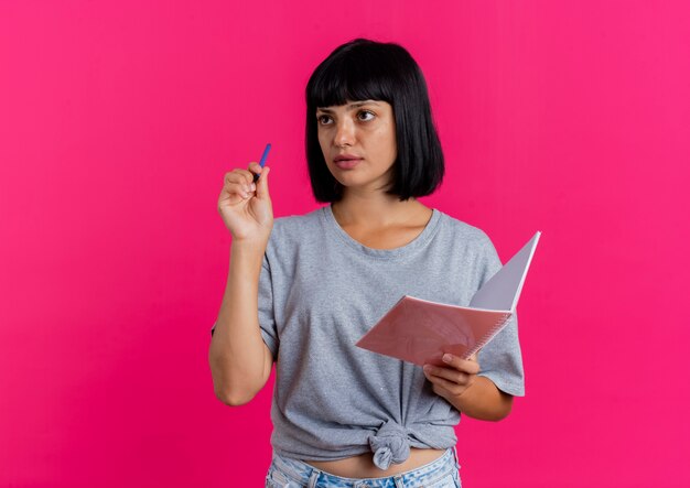 Schönes junges brünettes kaukasisches Mädchen hält Stift und Notizbuch, die Seite betrachten