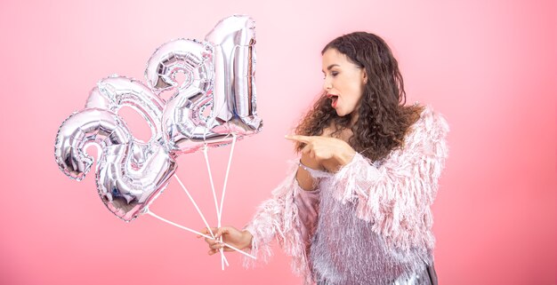 Schönes junges brünettes Mädchen mit lockigem Haar festlich gekleidet, das emotional auf einem rosa Hintergrund mit warmem Licht mit silbernen Luftballons für das Neujahrskonzept aufwirft