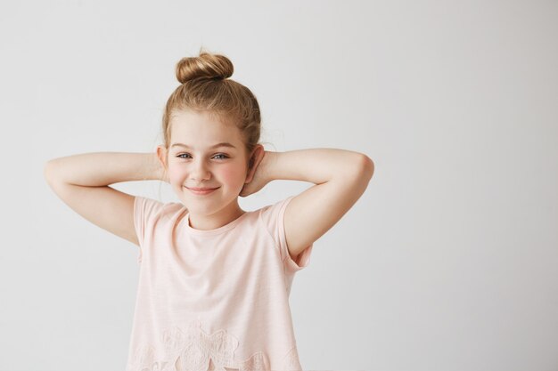 Schönes junges Fräulein mit blauen Augen und hellem Haar im rosa T-Shirt, das mit niedlicher Brötchenfrisur und Händen hinter Kopf für Familienfoto aufwirft.