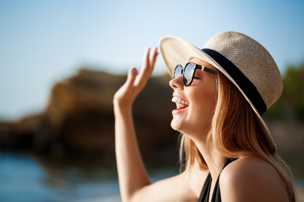 Schönes junges fröhliches Mädchen, das Brille und Hut trägt, ruht am Morgenstrand