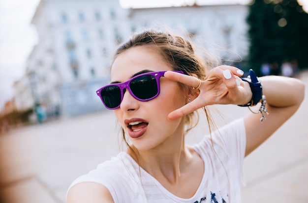 Schönes junges Mädchen in der Sonnenbrille, die zwei Finger, Friedensgeste zeigt