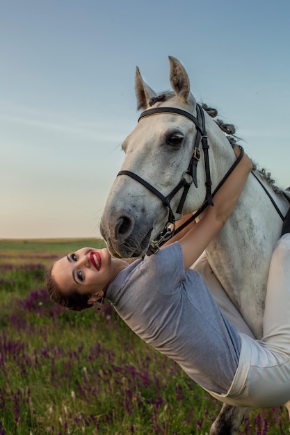 Schönes junges Mädchen lächelt ihr Pferd an, das einen einheitlichen Wettbewerb anzieht: Porträt im Freien bei Sonnenuntergang. Pflege von Tieren, Liebe und Freundschaftskonzept.