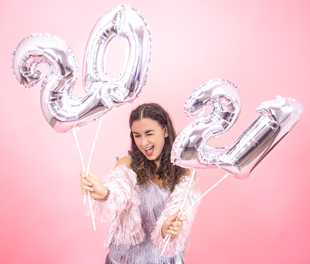 Schönes junges Mädchen mit einem Lächeln in einem festlichen Outfit auf einem rosa Studiohintergrund, der silberne Luftballons für das Neujahrskonzept hält