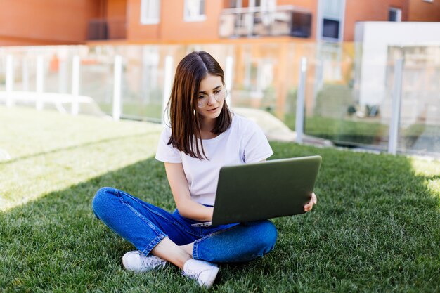 Schönes junges Mädchen mit Laptop draußen sitzend auf dem Gras