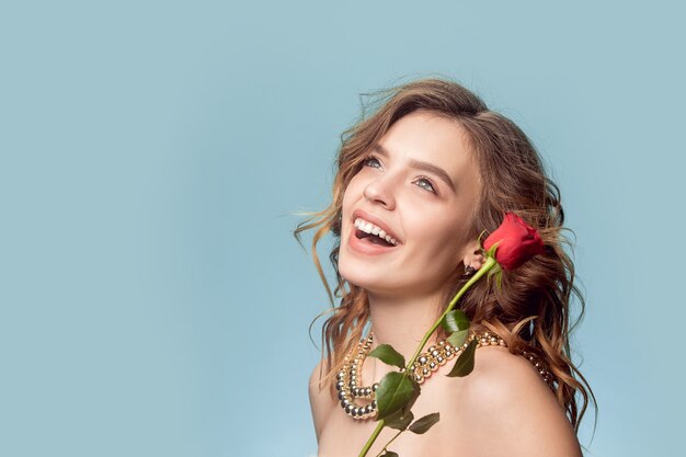 Schönes junges Mädchen mit rotem Rosen- und Perlenschmuck - Ohrringe, Armband, Halskette an der blauen Wand.